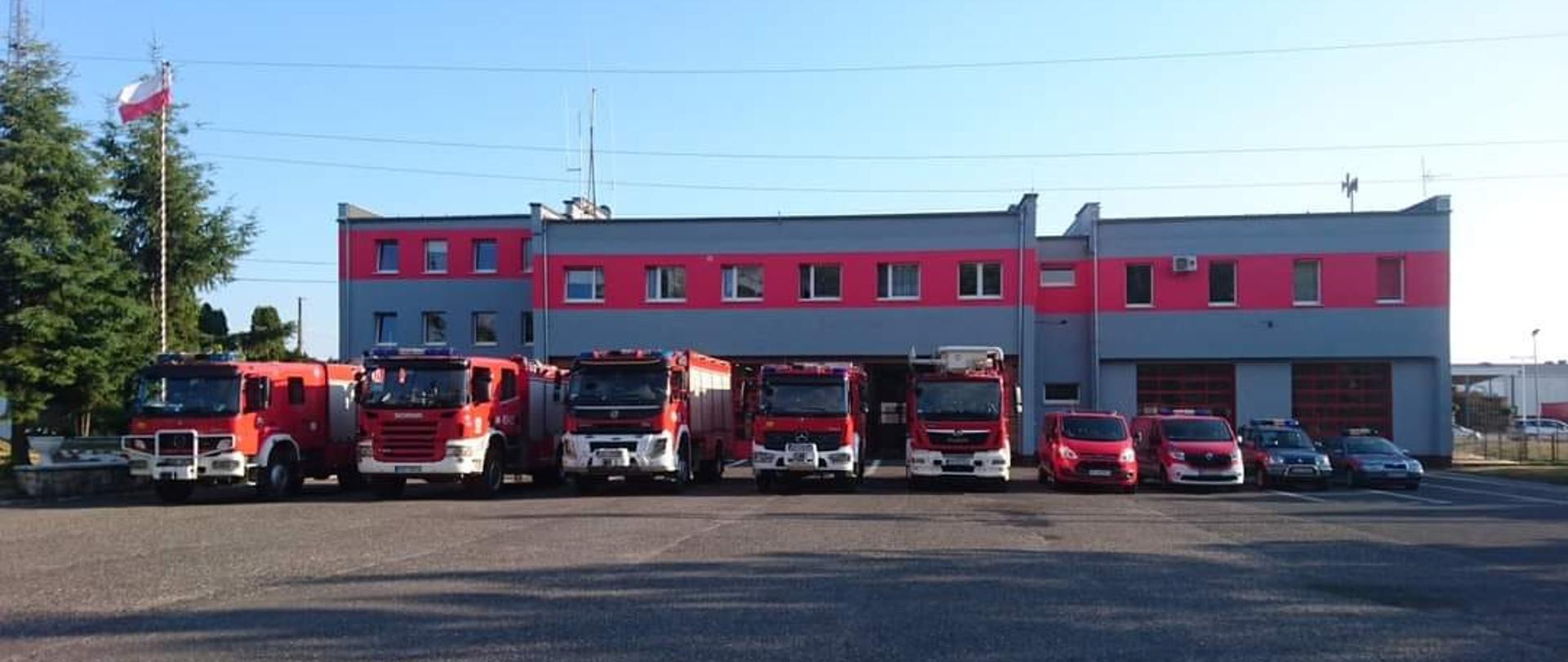 Zdjęcie przedstawia samochody Komendy Powiatowej PSP w Strzelcach Opolskich stojące w rzędzie przed siedzibą Komendy Powiatowej PSP w Strzelcach Opolskich. Pojazdy stoją na wyasfaltowanym placu wyjazdowym. Budynek w kolorze popielatym z czerwonym poziomym pasem na wysokości okien 1 piętra w 80% wysokości budynku. Po lewej stronie kadru flaga Polski na maszcie flagowym pomalowanym w biało-czerwone skośne pasy. Za masztem flagowym dwa drzewa iglaste.