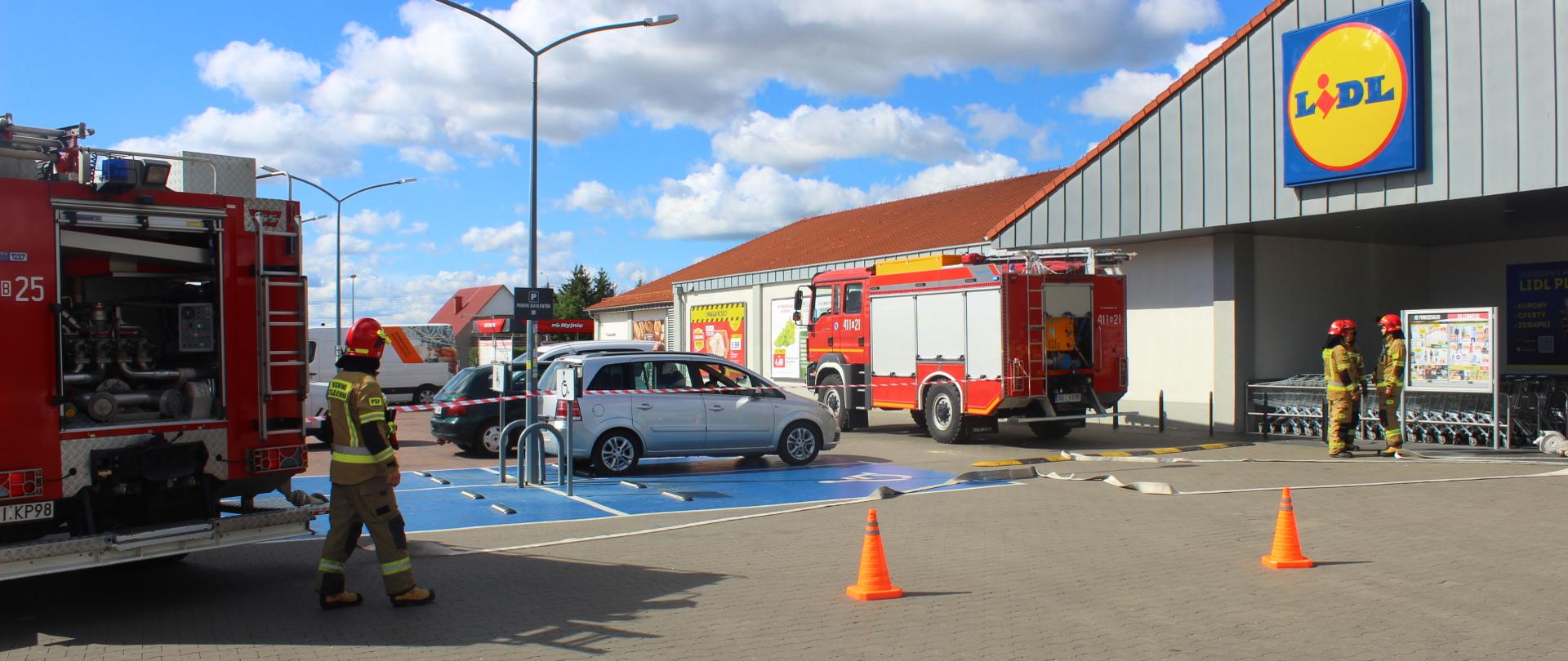 Zdjęcie przedstawia strażaków i dwa samochody ratowniczo-gaśnicze przed budynkiem sklepu lidl 