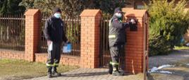 Na zdjęciu druh Ochotniczej Straży Pożarnej ubrany w umundurowanie bojowe wkładający ulotkę do skrzynki na listy zawieszonej na ogrodzeniu posesji.