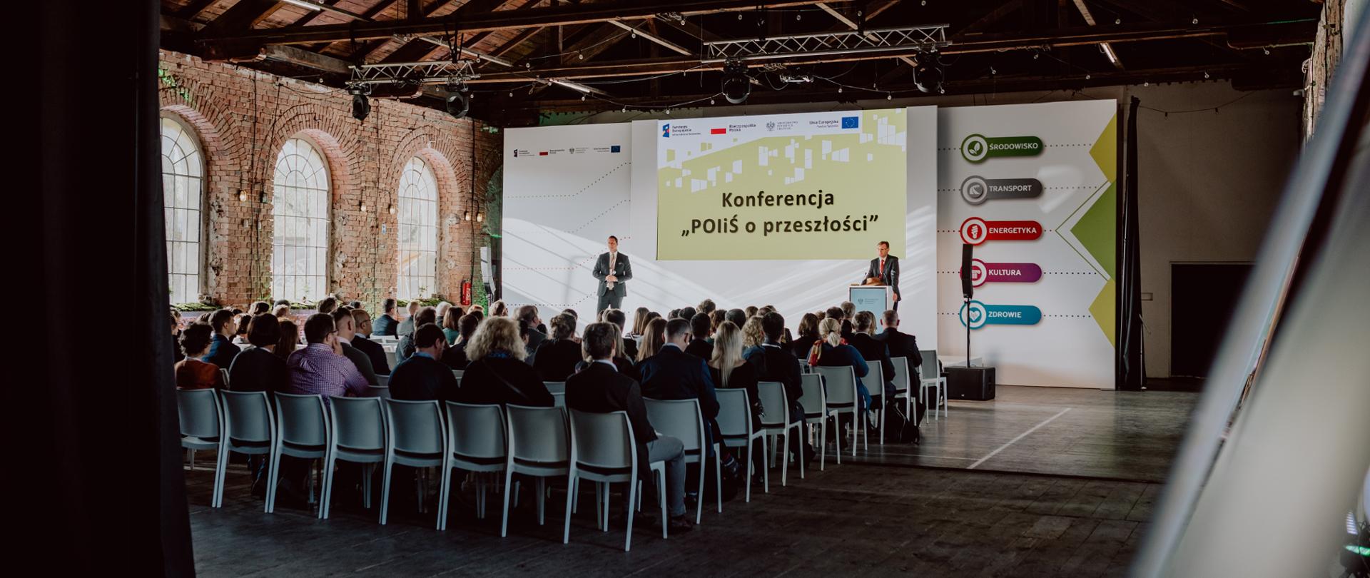 Zdjęcie przedstawia salę, na której na krzesłach siedzi grupa osób, przy mównicy stoi dyrektor Jarosław Orliński, a obok niego prowadzący. Za nimi wyświetlany jest napis "Konferencja POiŚ o przeszłości" 