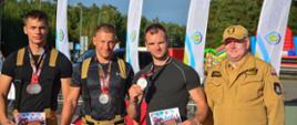 Trzech strażaków z medalami i dyplomami stoi obok przedstawiciela Państwowej Straży Pożarnej podczas zawodów w Jarosławcu.
