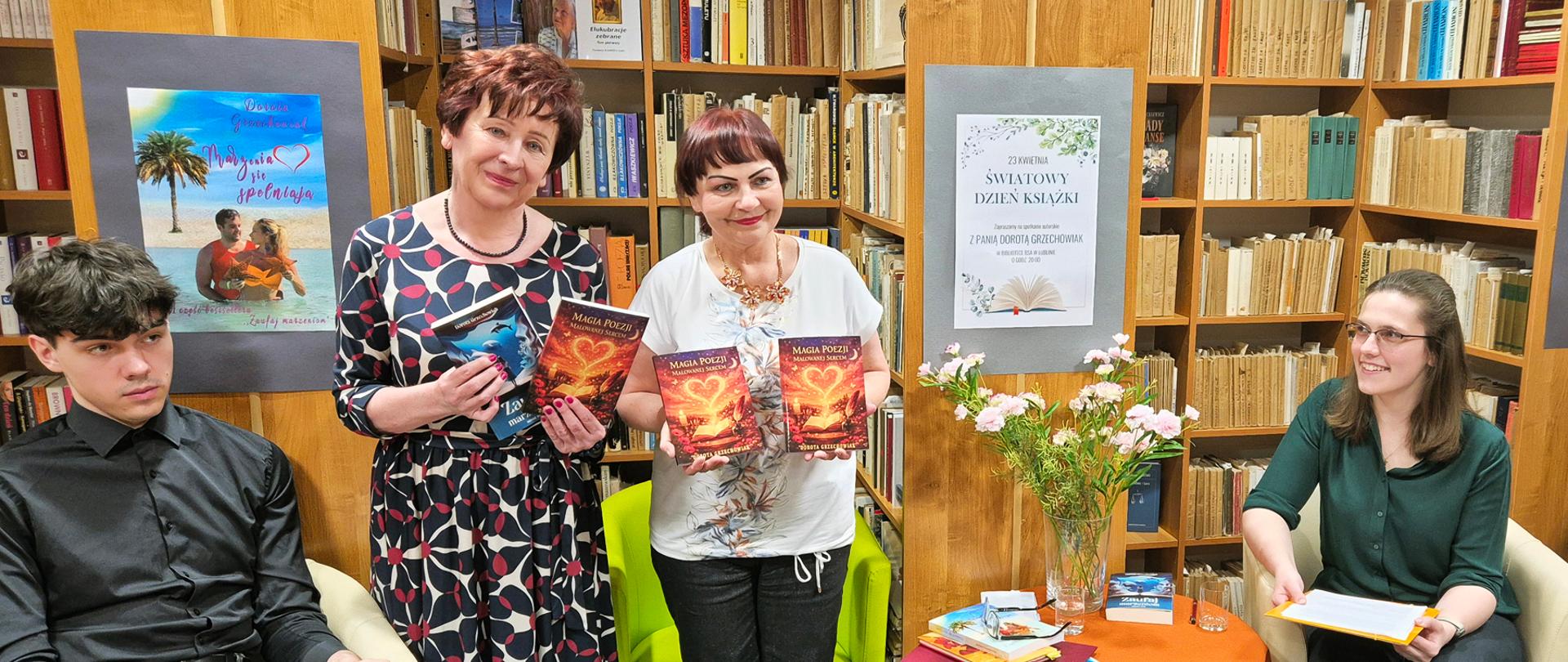 W pomieszczeniu biblioteki, na tle ściany z regałami wypełnionymi książkami stoją dwie kobiety po środku, trzymają w dłoniach książki. Po lewej siedzi chłopiec w fotelu , z po prawej siedzi dziewczyna w fotelu. Pomiędzy nią, a kobietą z książkami stoi stolik z bukietem kwiatów i leżącymi książkami.