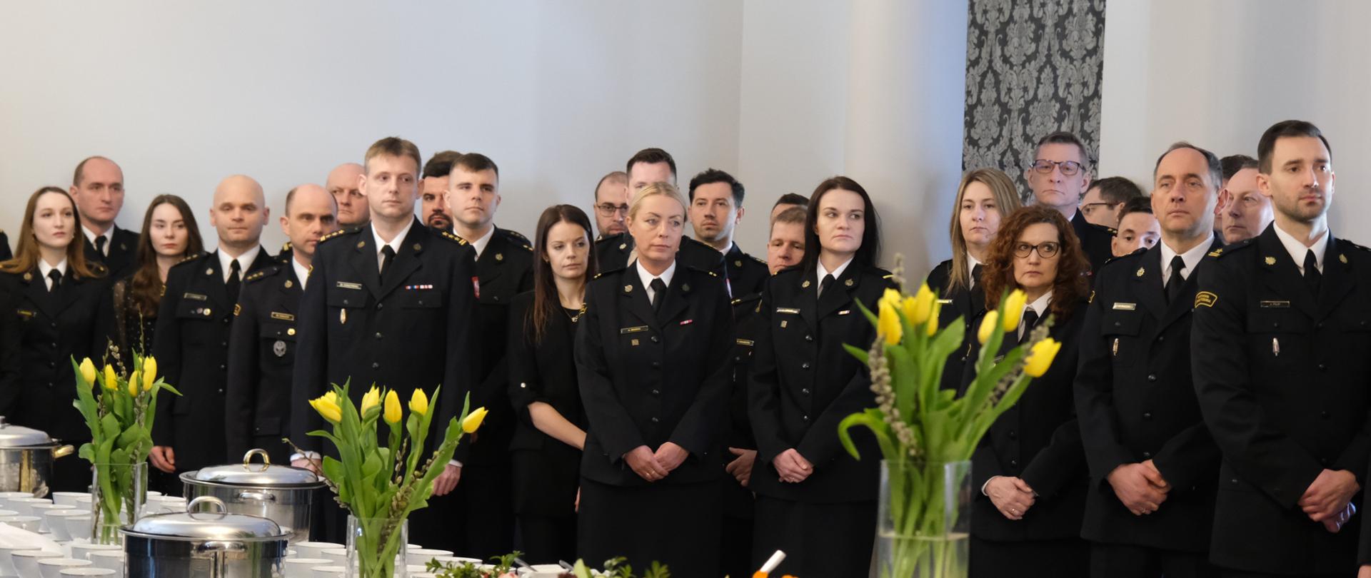 Pracownicy cywilni i funkcjonariusze Komendy Głównej PSP stoją w w sali słuchając życzeń komendanta głównego PSP podczas spotkania Wielkanocnego. Na pierwszym planie widoczne stoją ce na stołach bukiety żółtych tulipanów oraz gałązki wierzby