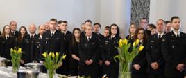 Pracownicy cywilni i funkcjonariusze Komendy Głównej PSP stoją w w sali słuchając życzeń komendanta głównego PSP podczas spotkania Wielkanocnego. Na pierwszym planie widoczne stoją ce na stołach bukiety żółtych tulipanów oraz gałązki wierzby