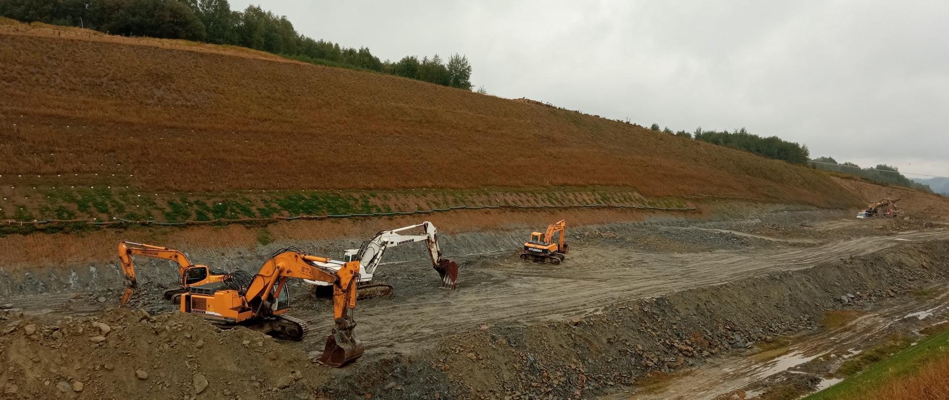 Prace ziemne na trasie główne na odc. S19 Babica - Jawornik. Widać korytarz drogi i wykop. W wykopie widać tony ziemi i pracujące koparki oraz kruszarki. Jest pogodnie.