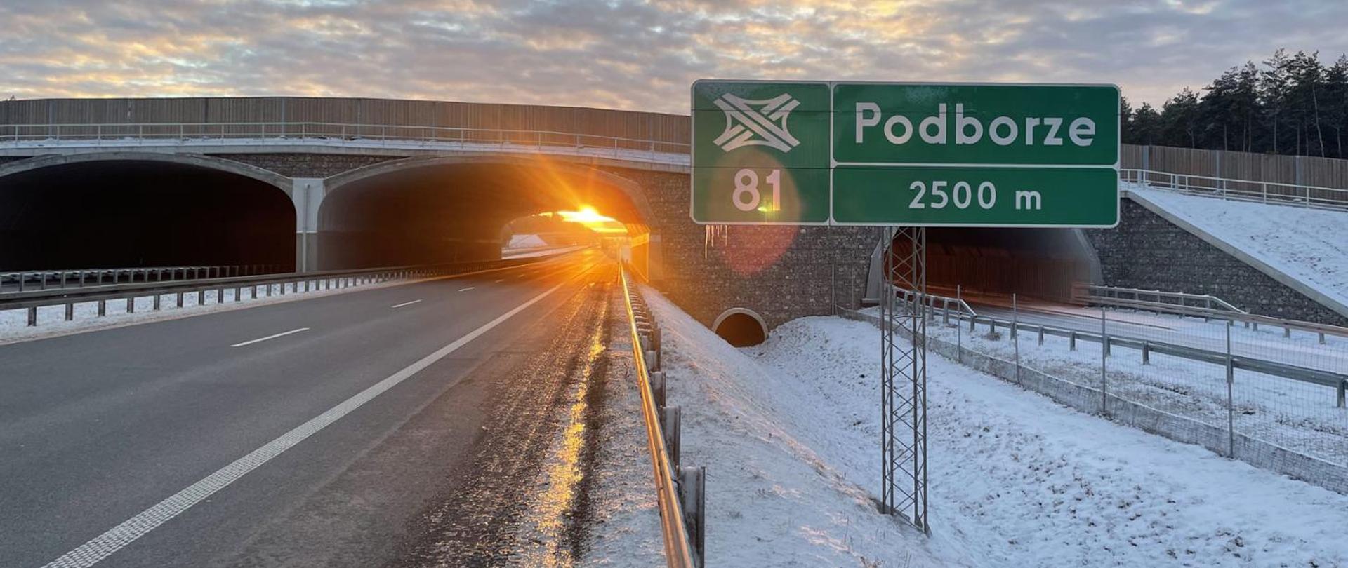 Droga ekspresowa biegnąca aż po horyzont. Na pierwszym planie tablica informująca o najbliższym węźle drogowym Podborze, za nią przejście dla zwierząt nad drogą, zachodzące słońce.