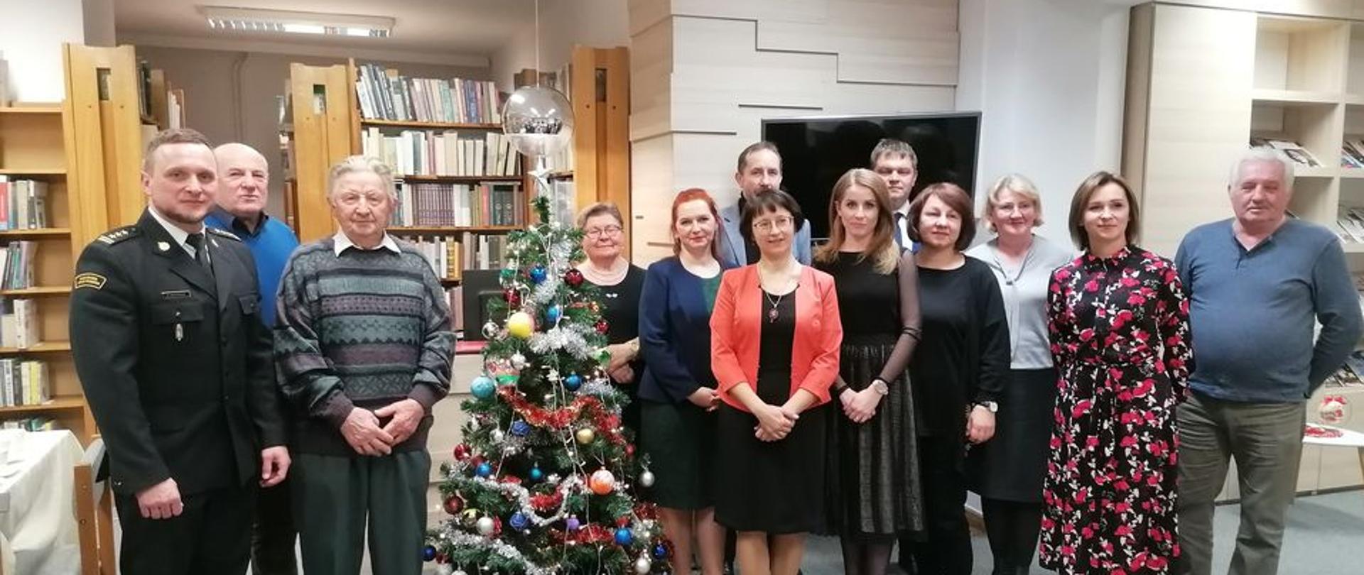 W pomieszczeniu obok choinki stoi kilkanaście osób.