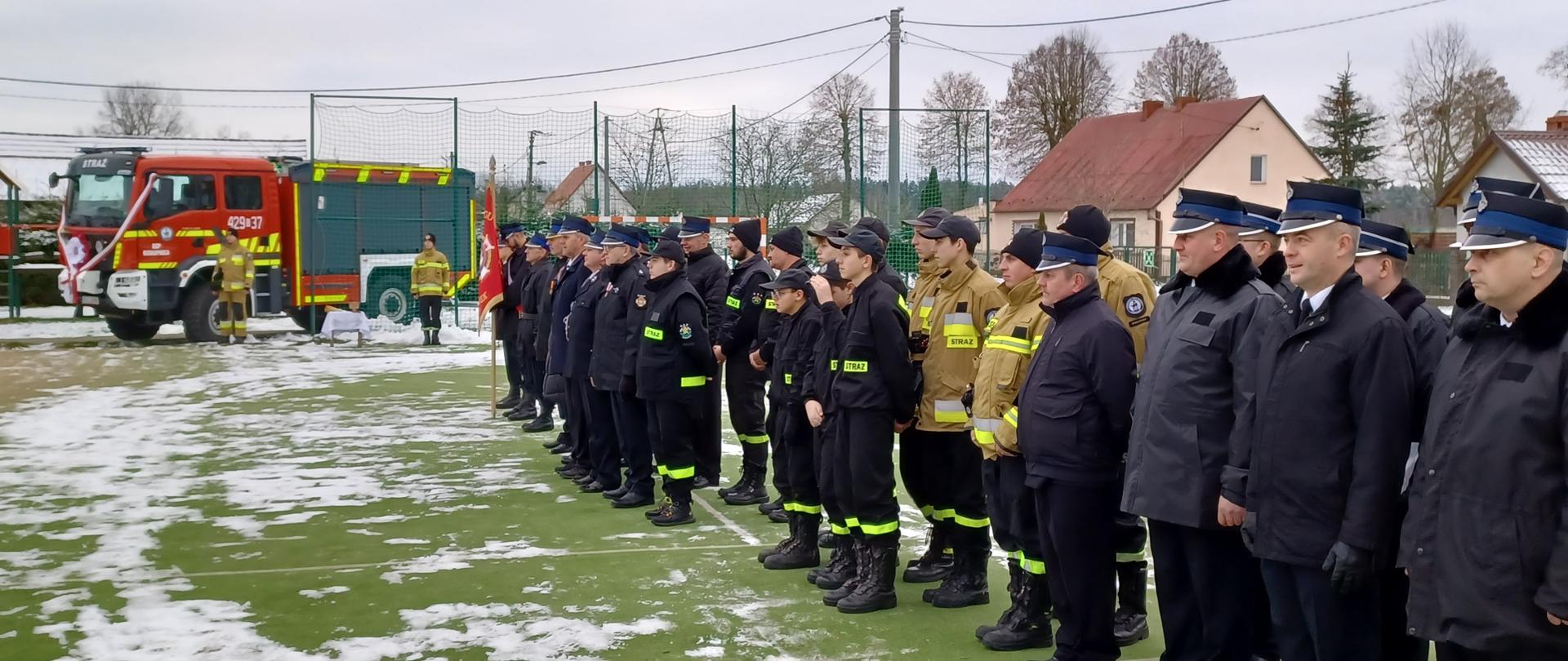 Strażacy stoją w dwuszeregu na boisku, w tle widać nowy samochód ratowniczo gaśniczy