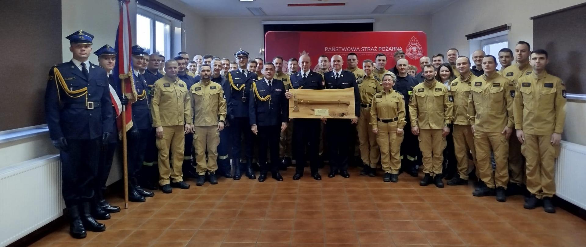 Na zdjęciu znajduje się duża grupa osób w umundurowaniu służbowym, ustawiona w kilku równych rzędach w przestronnym pomieszczeniu. Osoby po lewej stronie kadru mają na sobie ciemne mundury galowe, a jedna z nich stoi przy poczcie sztandarowym z flagą ustawioną na podstawie. Większa część grupy nosi jasne, beżowe stroje służbowe.
Centralnie, na przednim planie, kilka osób w ciemnych mundurach trzyma drewnianą skrzynkę lub pamiątkowy przedmiot prezentowany oficjalnie przed resztą zgromadzonych. W tle znajduje się czerwone tło z widocznym napisem „PAŃSTWOWA STRAŻ POŻARNA”, co wskazuje, że scena ma związek z uroczystością służbową strażaków.
Pomieszczenie jest jasne, z białymi ścianami, dużymi oknami po lewej stronie, ciemnymi roletami i podłużnymi lampami sufitowymi. Podłoga jest wyłożona brązowymi płytkami. Całość sprawia wrażenie oficjalnego wydarzenia – prawdopodobnie pożegnania, wyróżnienia lub innej uroczystości związanej z formacją strażacką.