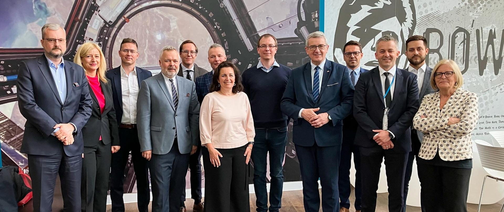 Na tle ściany w szare wzorki stoi duża grupa ludzi, pośrodku wiceminister Murdzek.