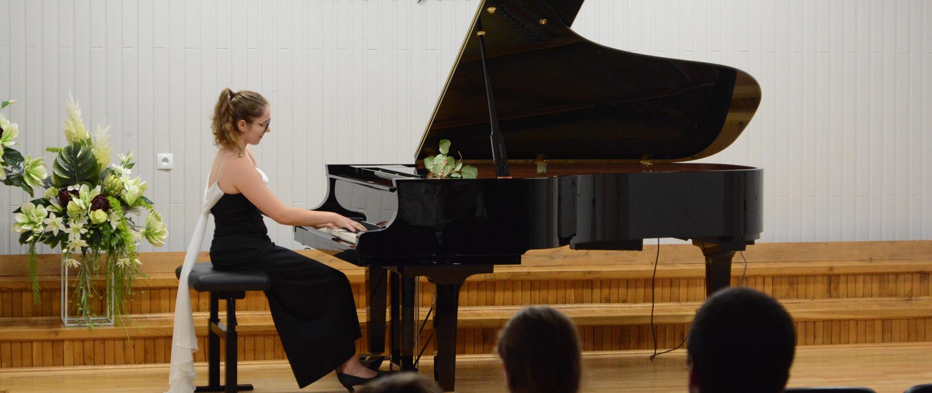 Na zdjęciu, w auli PSM widnieje Marianna Sereda, która koncertuje na fortepianie. Kolorystyka zdjęcia jest biało-brązowo-żółto-czerwono-zielono-czarna. 
