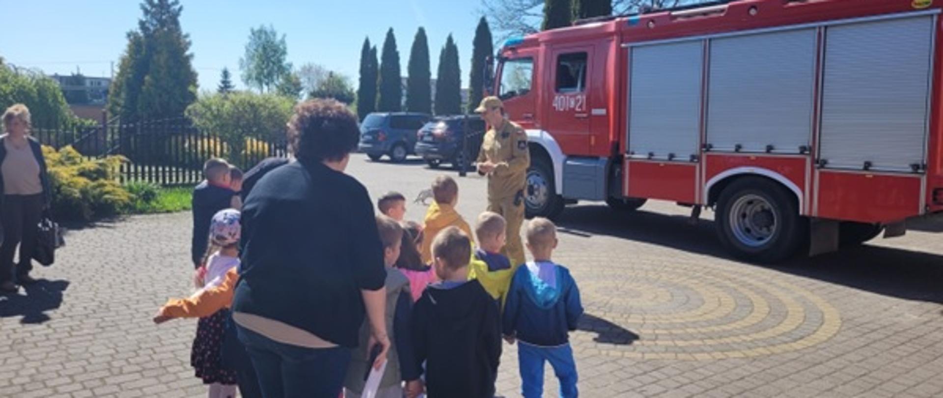 Na zdjęciu widać stojącą dzieci w różnokolorowych ubraniach oraz strażaka w piaskowym ubraniu koszarowym który opowiada co znajduje się w skrytkach samochodu pożarniczego który jest koloru czerwonego 