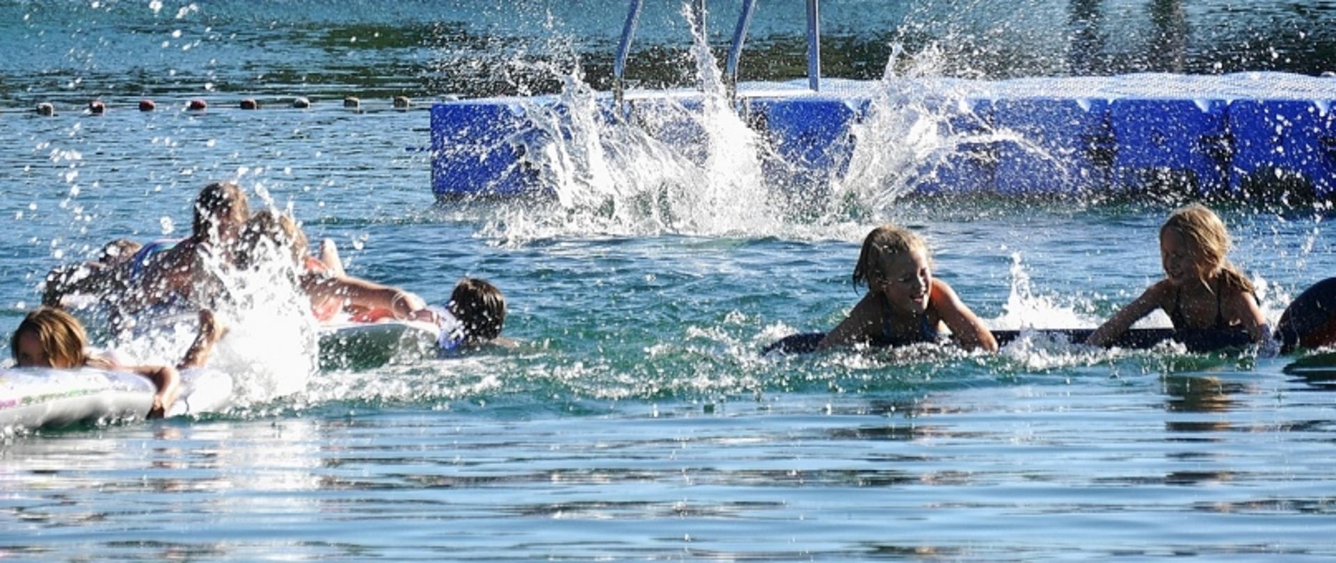 Grupa ludzi uczestnicząca w wodnej zabawie na tle spokojnego akwenu; widoczne rozpryski wody i niebieska platforma unosząca się na powierzchni.