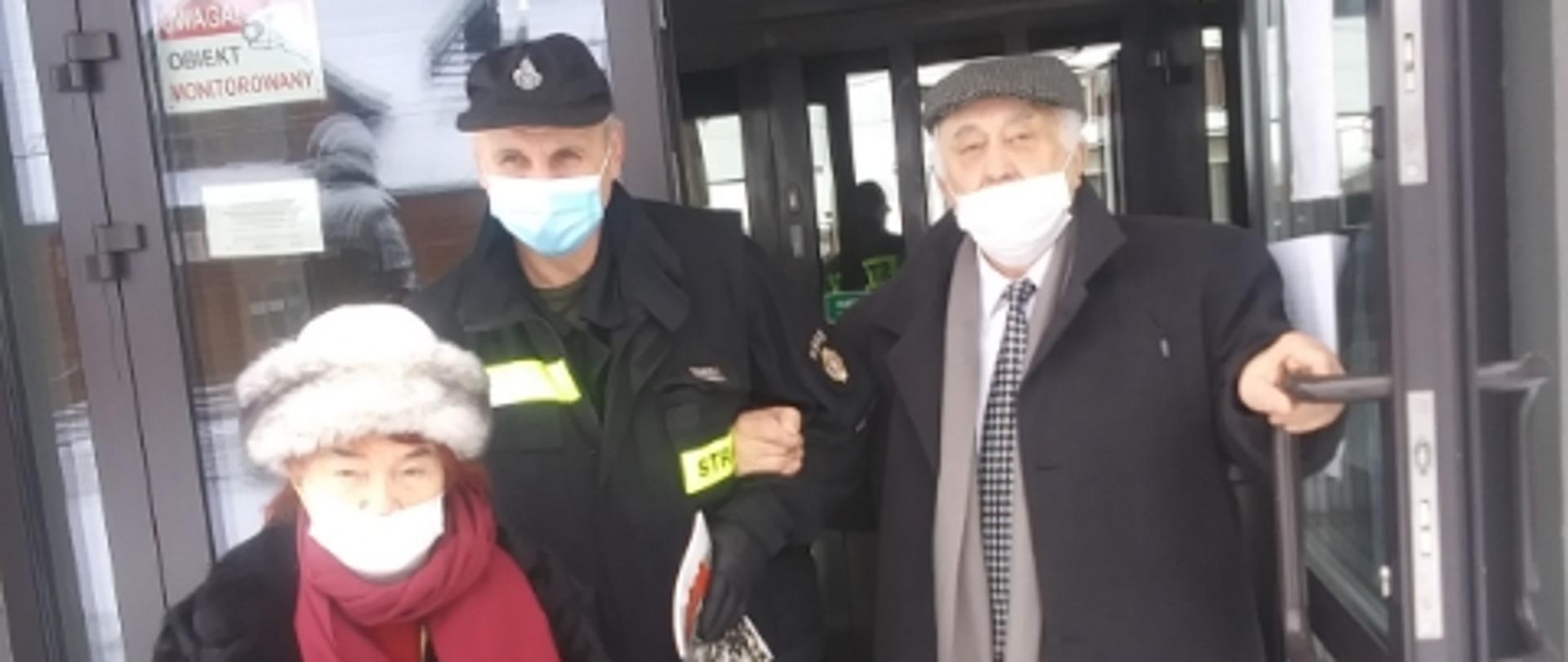 Zdjęcie przedstawia strażaka z Ochotniczej Straży Pożarnej w Kołakach Kościelnych w ubraniu koszarowym pomagającemu dwóm seniorom wychodzącym z pomieszczenia punku szczepień przeciwko COVID-19.