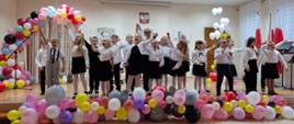 Zdjęcie ukazuje grupę dzieci wykonującą piosenkę na udekorowanej kolorowymi balonami scenie. Dzieci są radosne i uśmiechnięte. Ubrane są w białe bluzki i ciemne spódnice/spodnie. Za dziećmi widoczne jest godło Polski