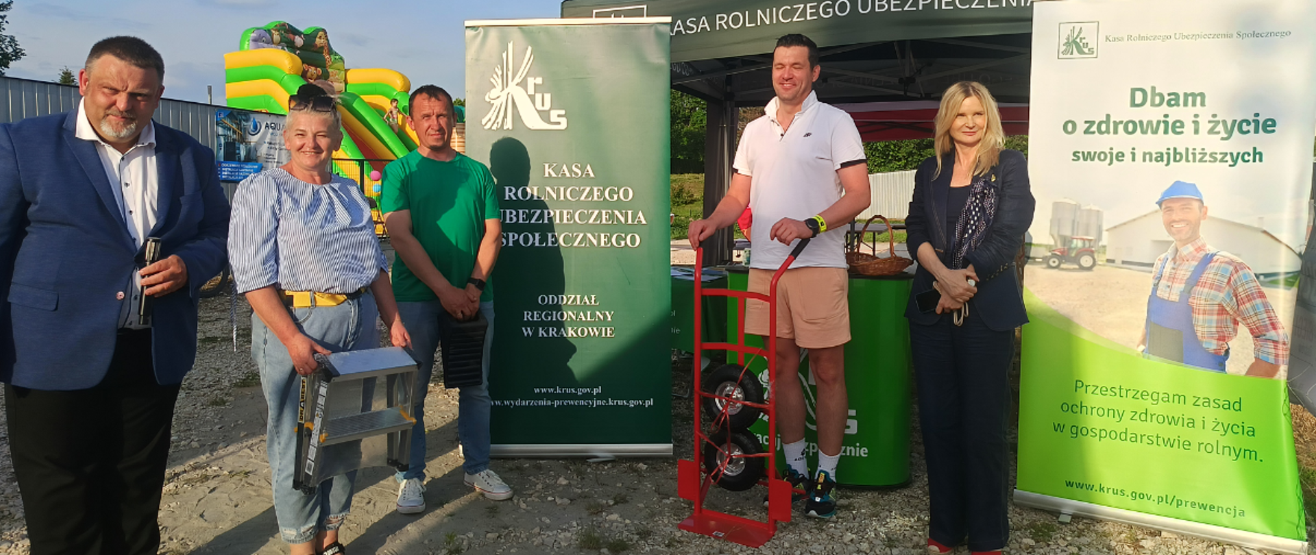 Na placu w Szreniawie przy namiocie ze stoiskiem informacyjno-prewencyjnym KRUS stoi pięć osób – dwie kobiety i trzech mężczyzn. Jedna z kobiet trzyma w rękach drabinkę, stojący po prawej mężczyzna trzyma ręczny wózek magazynowy. Wszyscy uśmiechnięci pozują do zdjęcia. Z tyłu widoczna trybunka reklamowa z logo KRUS, materiały prewencyjne, dwa banery.