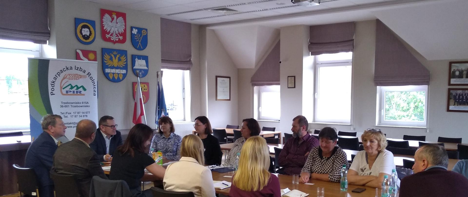 1. Grupa ludzi siedząca w dużym pomieszczeniu przy stole. W tle stoi baner. W ścianach znajduje się duża ilość okien oraz wiszą fotografie.