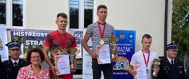 na podium stoją trzej sportowcy, po bokach umundurowani strażacy, w tle jasny budynek