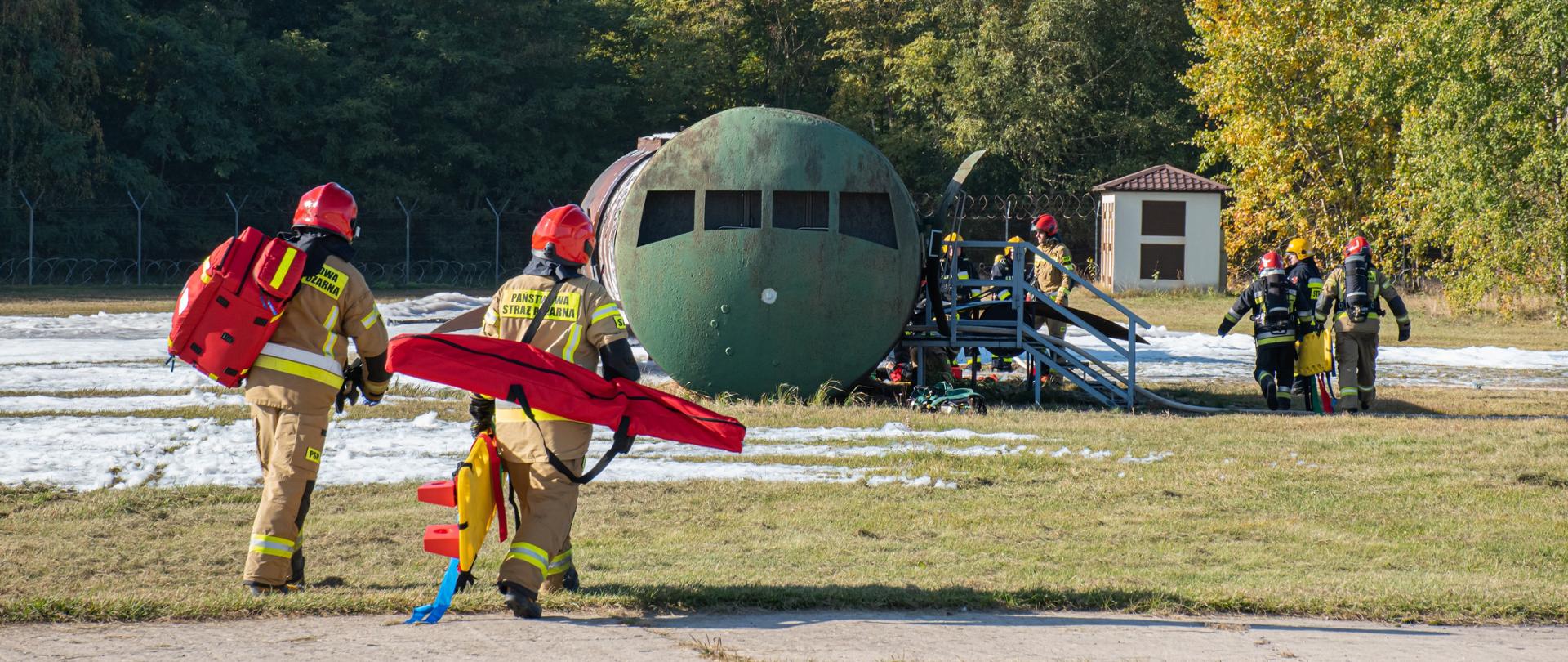 Strażacy z torbą medyczna i noszami idą w kierunku wraku samolotu (symulator wypadku lotniczego)