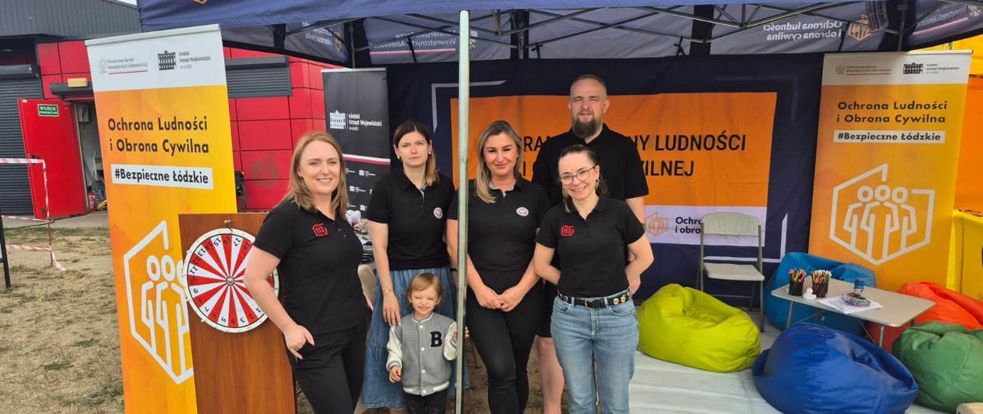 Pięcioro pracowników Łódzkiego Urzędu Wojewódzkiego pozują do zdjęcia w namiocie. W tle plakaty Programu Ochrony Ludności i Obrony Cywilnej oraz pufy dla dzieci i kolorowanki.