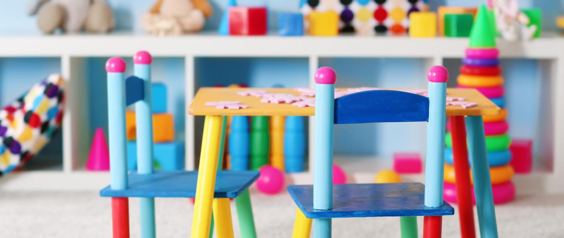 Dziecięcy pokój z zabawkami. Na pierwszym planie dwa kolorowe krzesełka przy stoliku. Na drugim planie na białej półce widać zabawki - kocki, pluszaki.