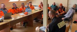 Strażak na Sali wykładowej wykłada prelekcję na temat pierwszej pomocy. Na Sali pracownicy firmy fb serwis w pomarańczowych ubraniach.