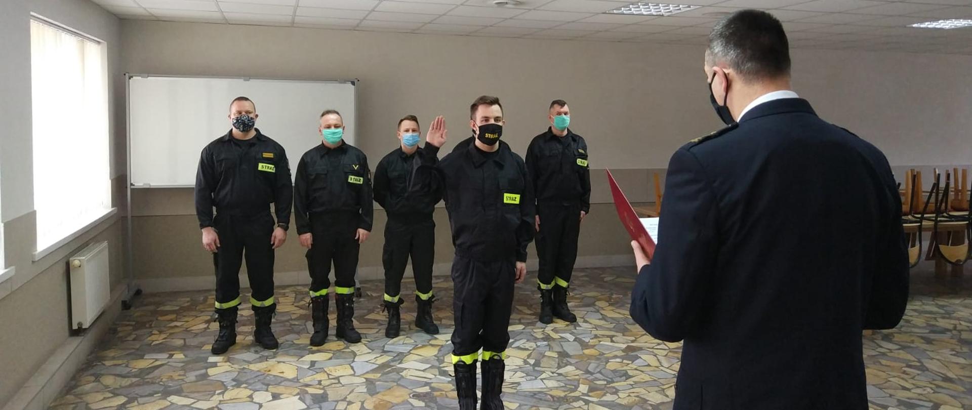 Zdjęcie przedstawia złożenie ślubowania przez nowo zatrudnionego funkcjonariusza w obecności Komendanta Powiatowego PSP w Oleśnicy. Ślubowanie odbywa się w świetlicy JRG w Sycowie. W tle znajduję się zmiana służbowa.