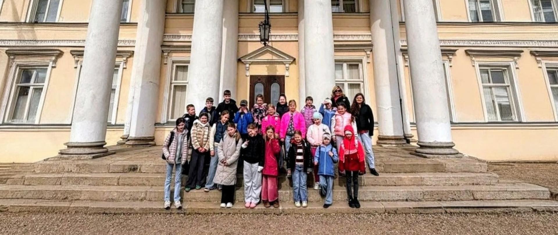 kilkunastoosobowa grupa osób stojąca na schodach przed zabytkowym pałacem, za nimi wysokie masywne białe kolumny 