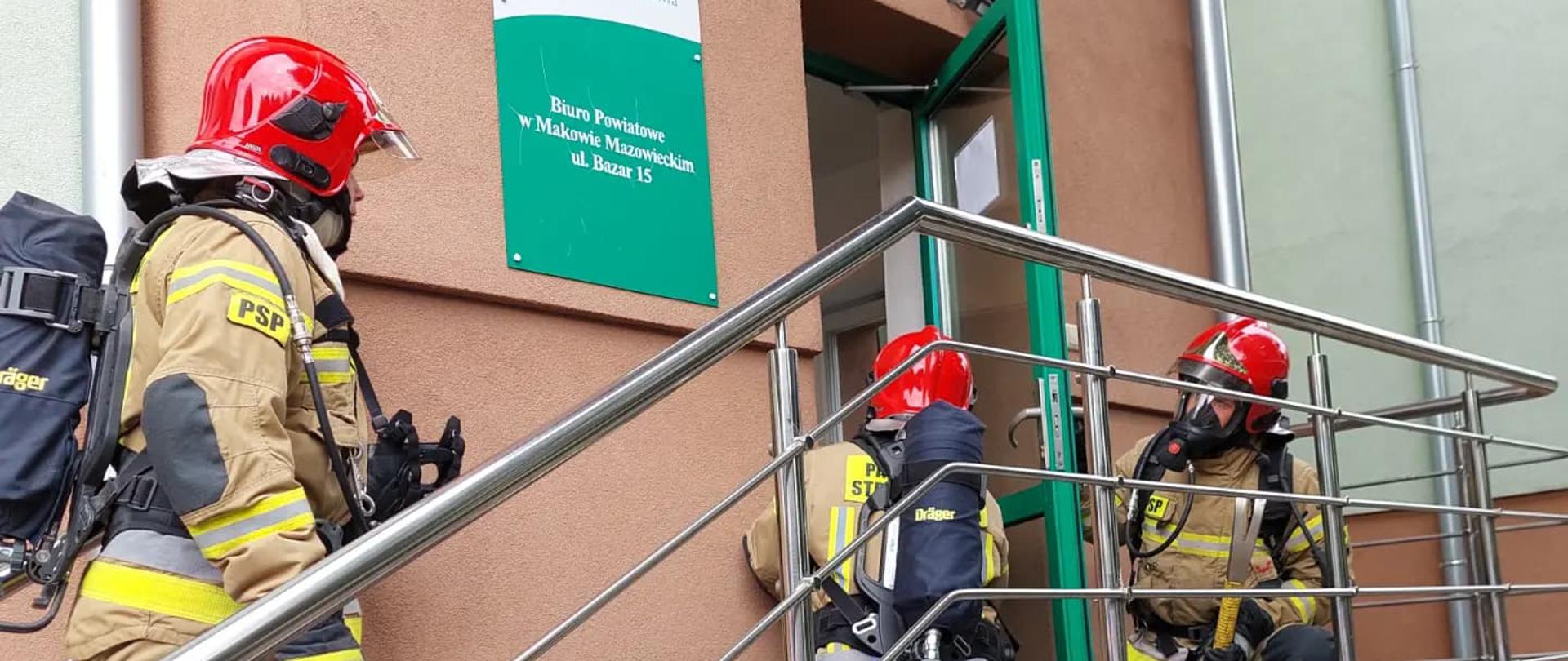 Trzech strażaków stoi na schodach przed wejściem do budynku. Jeden z nich otwiera drzwi.