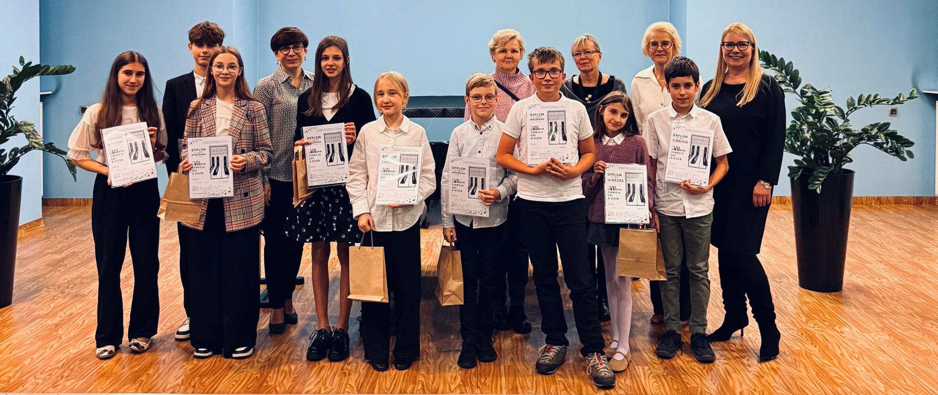Na kolorowym zdjęciu widać grupę osób stojących na scenie w jasnoniebieskiej sali. W pierwszym rzędzie znajduje się kilkoro dzieci i młodzieży, którzy trzymają w rękach dyplomy oraz papierowe torebki z nagrodami. Za nimi stoją dorośli, nauczyciele i organizatorzy wydarzenia. Wszyscy są uśmiechnięci i ubrani w stroje w kolorach odświętnych. Po bokach sceny stoją duże donice z zielonymi roślinami.