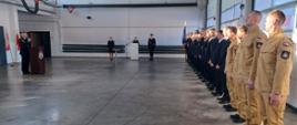 Zdjęcie zrobione w pomieszczeniu strażnicy. Na pierwszym planie po prawej stronie strażacy ustawieni w szeregu, w umundurowaniu służbowym oraz galowym, po lewej komendant powiatowy Państwowej Straży Pożarnej w Lublińcu st. bryg. Janusz Bula stojący za mównicą przemawia do mikrofonu. Na wprost trzech strażaków w postawie stojącej w umundurowaniu galowym, jedna osoba za mównicą.