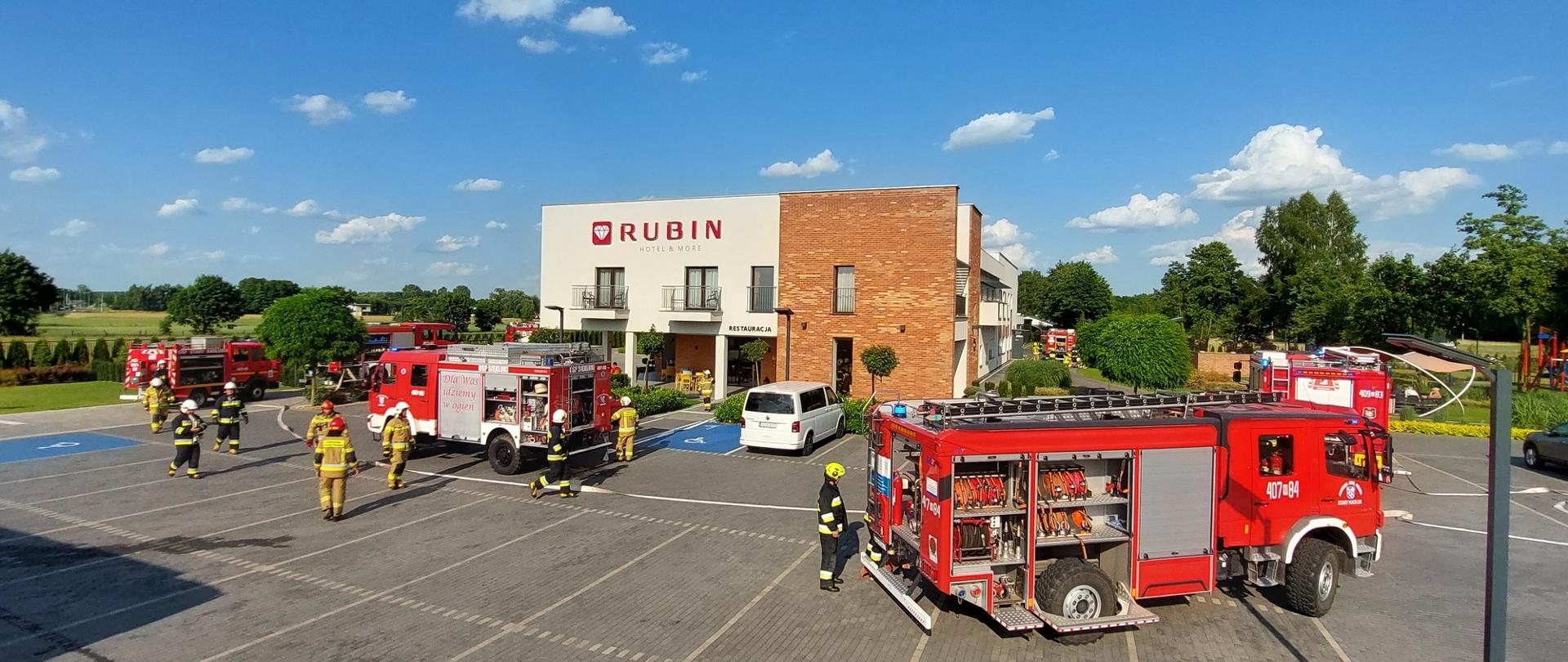 Siedem samochodów pożarniczych na parkingu przed hotelem RUBIN, strażacy w ubraniach specjalnych prowadzą działania ratowniczo-gaśnicze.