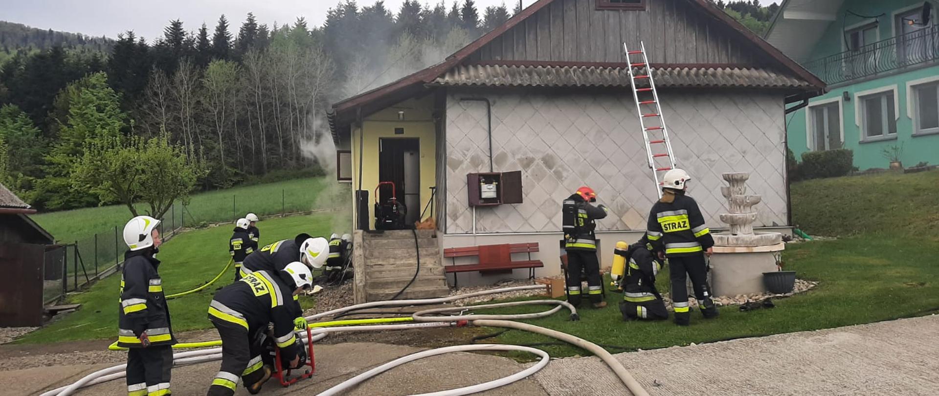Linie gaśnicze na zewnątrz budynku oraz strażacy prowadzący działania ratownicze. Z budynku unosi się dym.