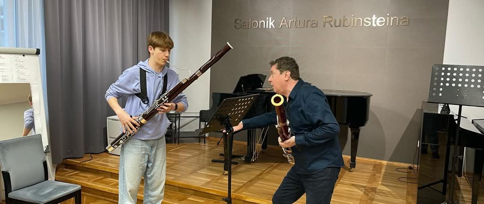 na zdjęciu na pierwszym planie uczeń w niebieskim ubraniu grający na fagocie po lewej stronie profesor w ręce trzymający fagot w tle fortepian