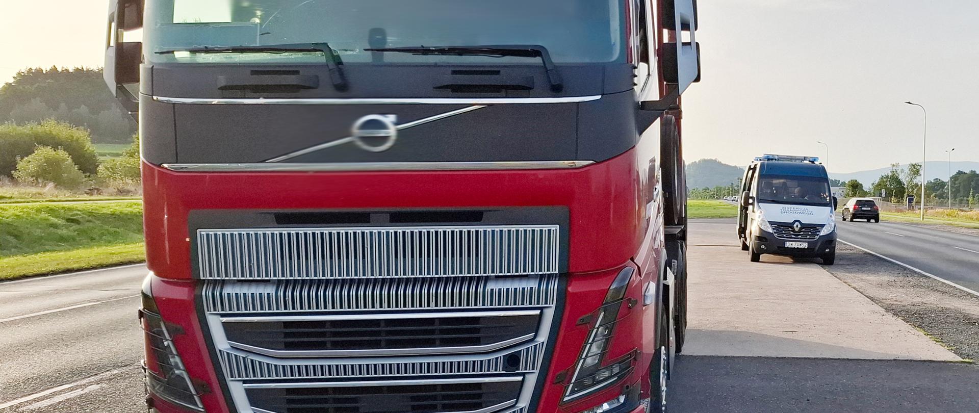 Na pierwszym planie przód pojazdu ciężarowego , za nim inspekcyjny furgon. W tle droga z jadącym samochodem osobowym i przydrożna infrastruktura.