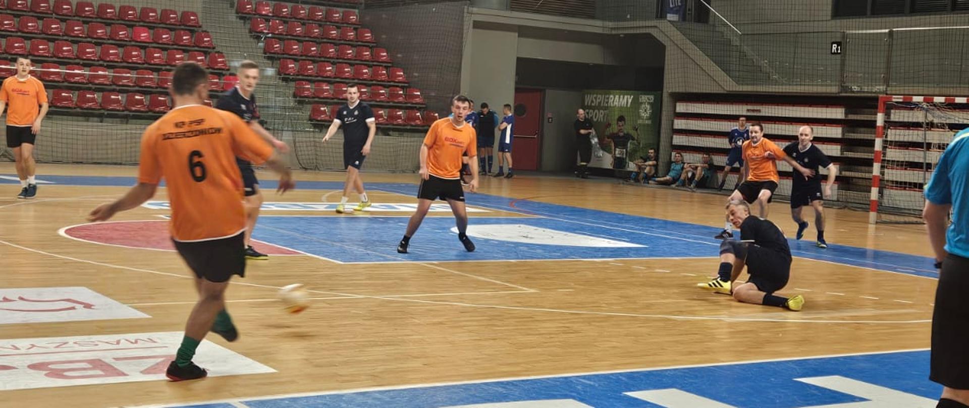 Zawodnicy w strojach granatowych oraz pomarańczowych grają w piłkę halową na sali