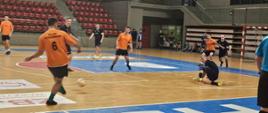 Zawodnicy w strojach granatowych oraz pomarańczowych grają w piłkę halową na sali