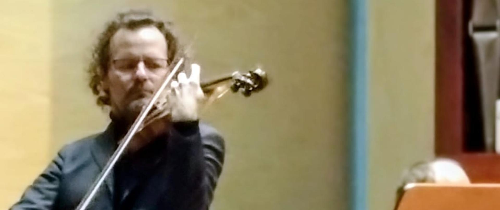 To jest pionowe, średnie ujęcie z koncertu lub występu muzycznego, skupiające się na muzyku grającym na skrzypcach. Główna Postać: W centrum kadru stoi dorosły mężczyzna z jasną skórą, w eleganckim, ciemnym garniturze i czarnym golfie. Ma kręcone, brązowe włosy i zarost. Jest uchwycony w trakcie dynamicznego grania na skrzypcach, trzymając instrument pod brodą, a smyczek w prawej ręce. Jego postawa jest skupiona i profesjonalna. Tło: Tło jest proste, składa się z gładkiej, jasnej ściany (prawdopodobnie w kolorze kremowym lub jasnego beżu). Widać fragmenty sceny: Po prawej stronie znajduje się wysoki, drewniany pulpit na nuty z dużą, otwartą partyturą. Za pulpitem, w głębi, siedzi inna, słabo widoczna postać (prawdopodobnie pianista) na czarnym taborecie, przed ciemnym fortepianem. W prawym górnym rogu widać fragment ciemnobrązowego elementu, być może części organów piszczałkowych lub innej konstrukcji. Nastrój: Oświetlenie jest ciepłe i jasne, co sugeruje formalną atmosferę sali koncertowej. Zdjęcie oddaje moment intensywnego wykonania muzycznego.