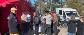Na zdjęciu znajduje się grupa młodych ludzi zgromadzonych wokół czerwonego namiotu. Na pierwszym planie stoi mężczyzna w mundurze strażackim z naszywką PSP (Państwowa Straż Pożarna) i flagą Polski na rękawie, uśmiechając się do kamery. Część osób czeka w kolejce do namiotu, inni rozmawiają w grupach. W tle widać białego busa, osobę na wózku inwalidzkim oraz strażacki sprzęt leżący na ziemi. Sceneria wskazuje na wydarzenie związane z edukacją przeciwpożarową lub ćwiczeniami służb ratunkowych na świeżym powietrzu, przy dobrej pogodzie.
