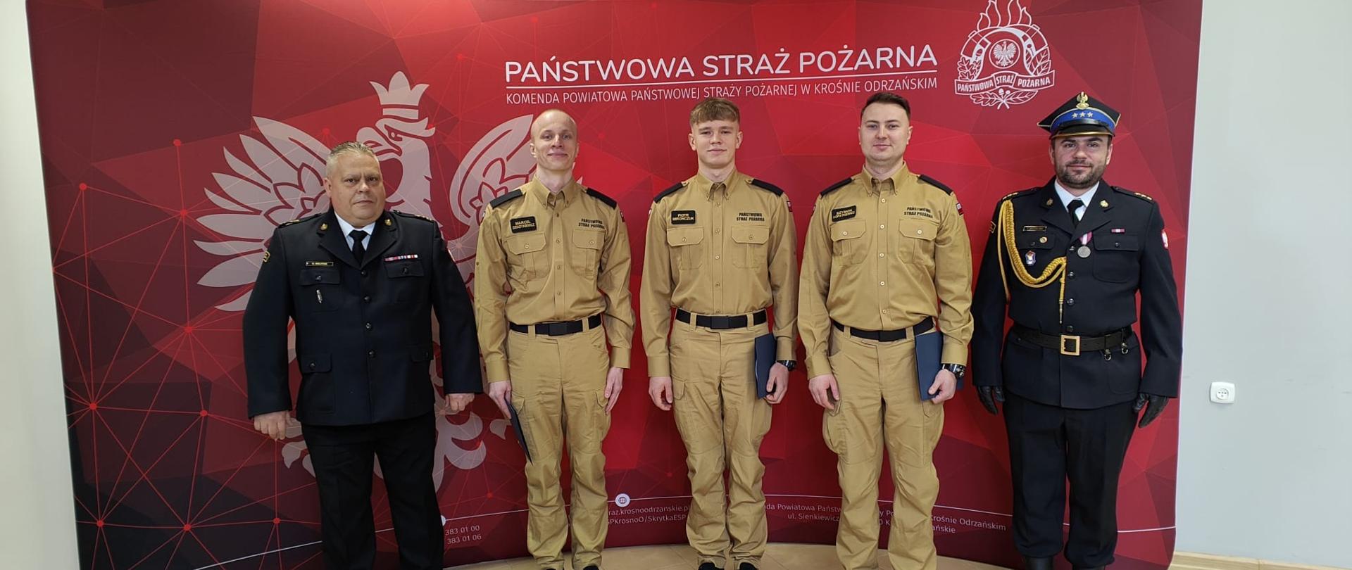 Grupowe zdjęcie pięciu strażaków wykonane na tle czerwonej ścianki Państwowej Straży Pożarnej w Krośnie Odrzańskim. Trzech strażaków w jasnym umundurowaniu stoi pomiędzy dwoma funkcjonariuszami w mundurach galowych.