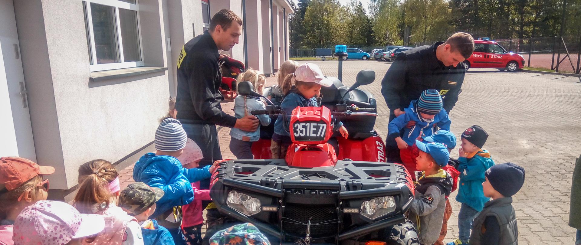 Zdjęcie przedstawia grupę dzieci podczas zapoznania ze sprzętem strażackim, na pojeździe typu quad na tle budynku.