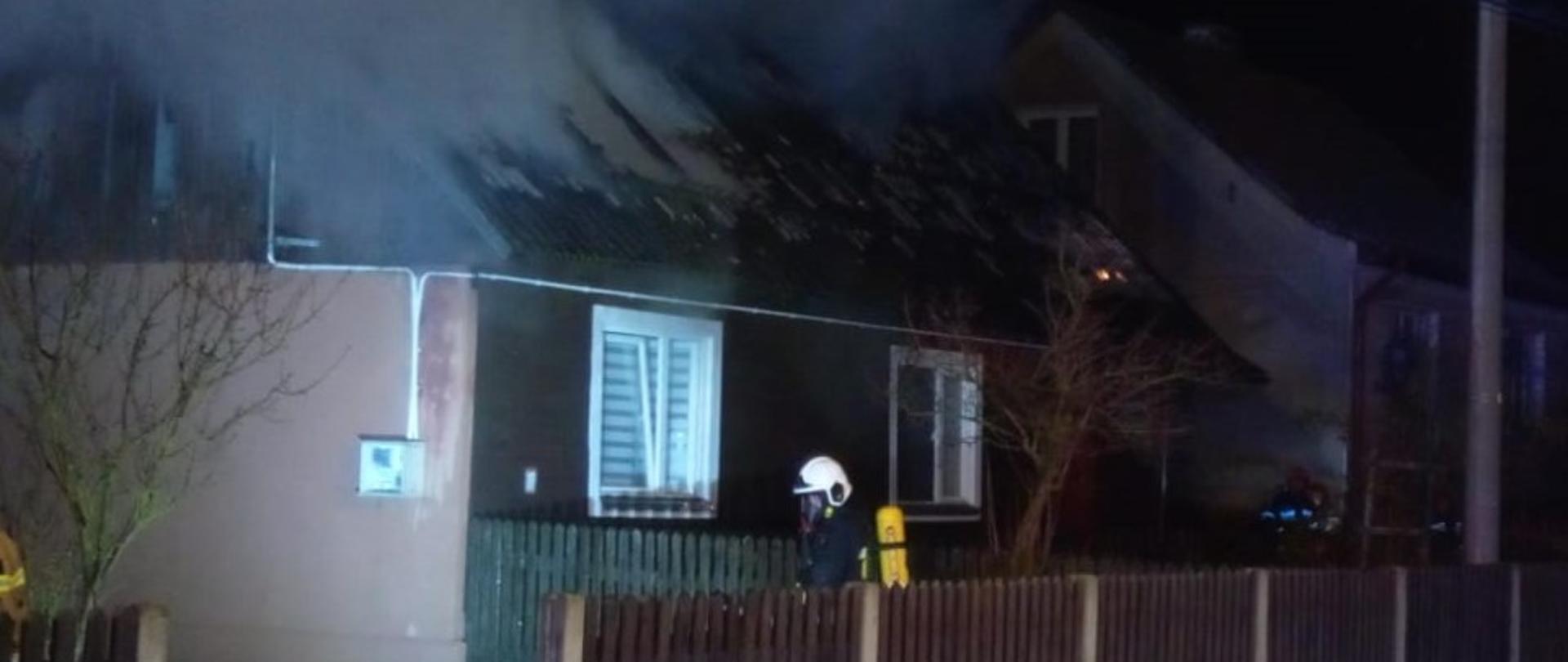 Zdjęcie przedstawia dom jednorodzinny, w którym paliło się poddasze nieużytkowe. Przed budynkiem widać strażaka w aparacie ochrony dróg oddechowych.