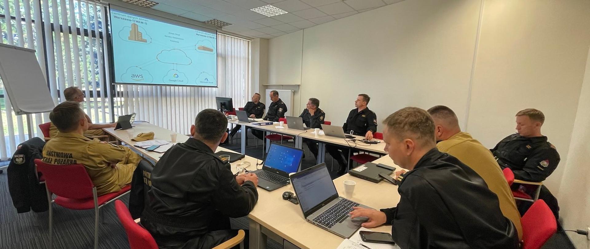 Grupa funkcjonariuszy w mundurach koloru czarnego i piaskowego siedzi przy stołach, przed nimi na stole laptopy a na froncie stołów ustawiony monitor, na którym wyświetlane są slajdy