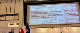 Mężczyzna w mundurze przemawia. Za nim wyświetlana jest prezentacja dotycząca Programu Ochrony Ludności i Obrony Cywilnej