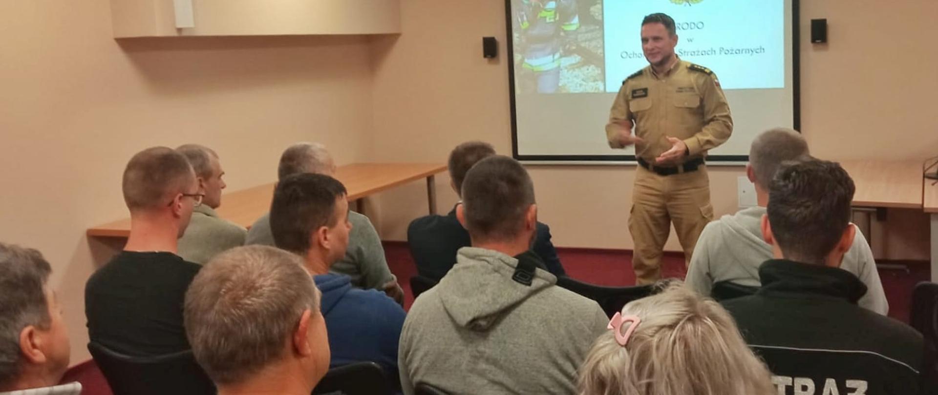 Na sali na krzesłach siedzi kilkadziesiąt osób. Przed nimi stoi strażak w piaskowym mundurze.