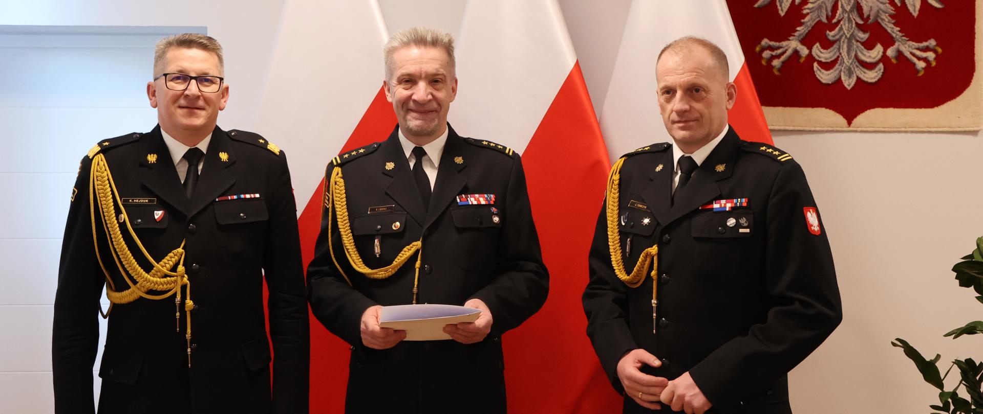 Zdjęcie pamiątkowe. Trzech mężczyzn w mundurach pozujący na tle flag narodowych i godła