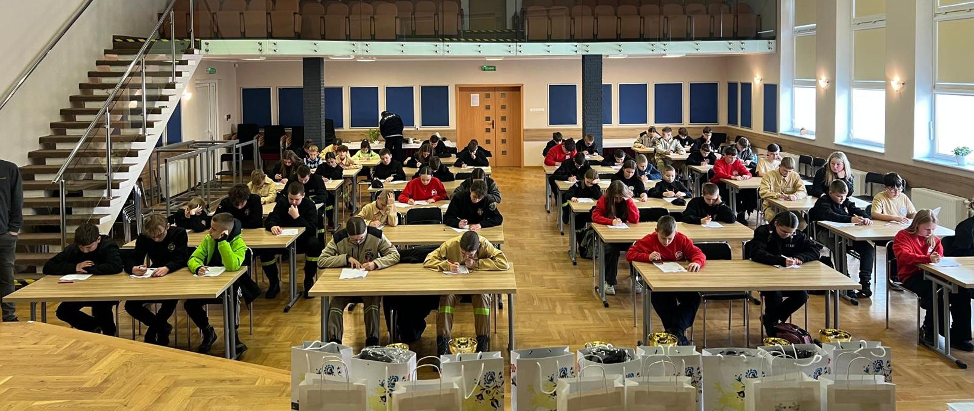 Na pierwszym planie nagrody rzeczowe oraz puchary dla najlepszych uczestników. Na dalszym planie dzieci i młodzież rozwiązują test wiedzy z zakresu pożarnictwa.