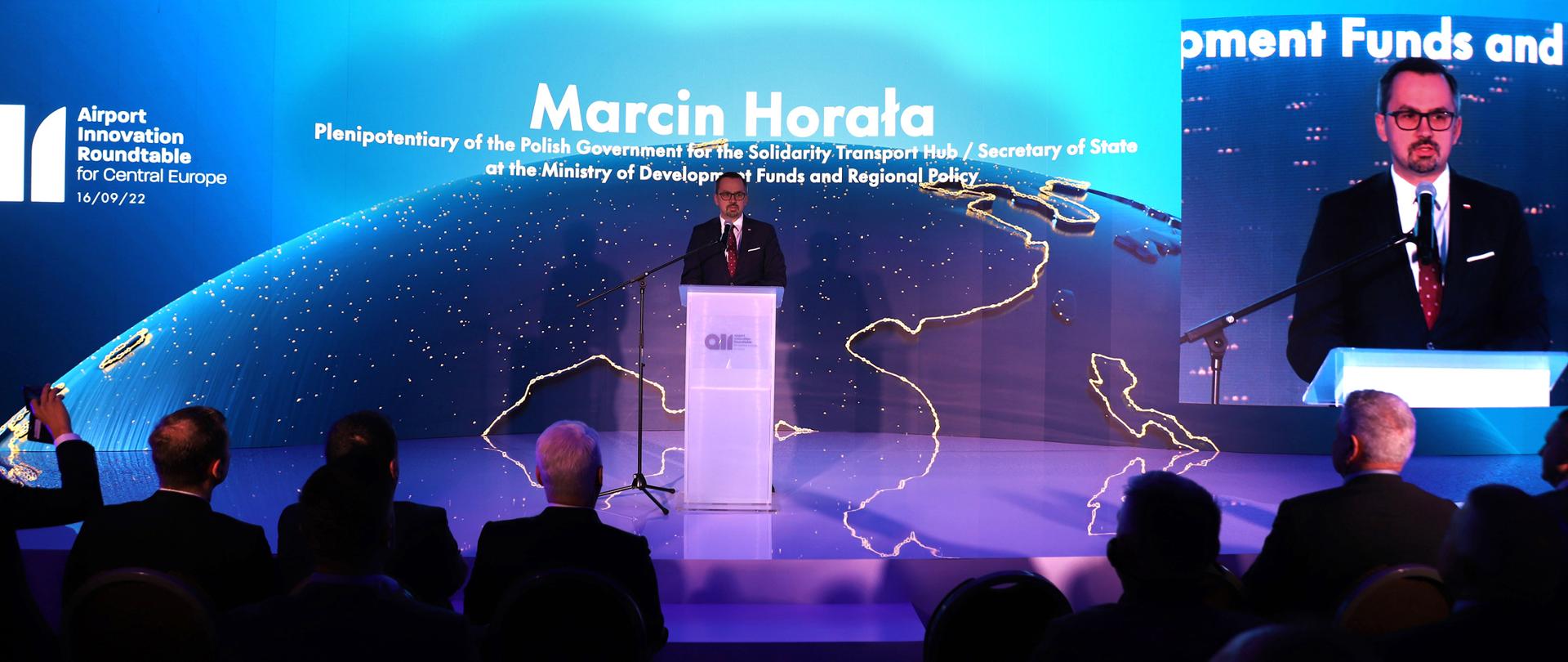 Minister Marcin Horała stoi na scenie przy podeście. Mówi do ludzi na widowni, którzy siedzą na przeciwko niego. 