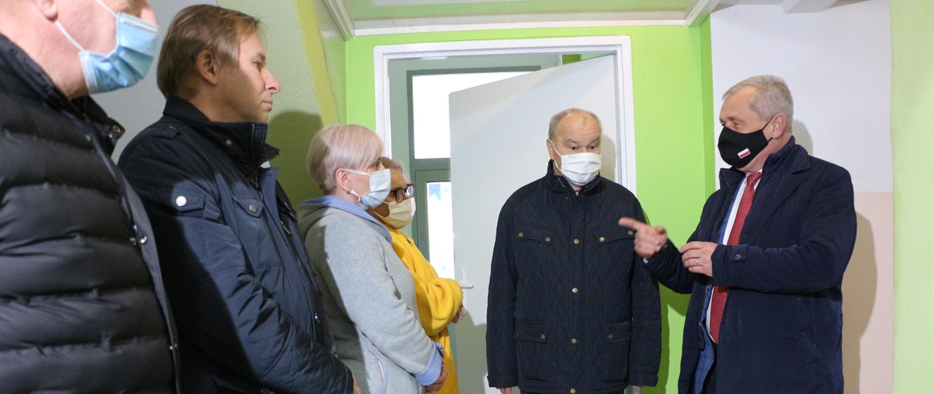 Na zdjęciu wojewoda opolski po prawej oraz - obok niego, dyrektor szpitala w Nysie. Panowie stoją na przeciw kadry medycznej. Rozmawiają na temat Covid-19. Wojewoda, mający na twarzy maskę ochronną, skierowany jest ciałem w kierunku rozmówców, ma uniesiony palec, którym wskazuje na rozmówców. Wszyscy stoją na korytarzu w szpitalu. 