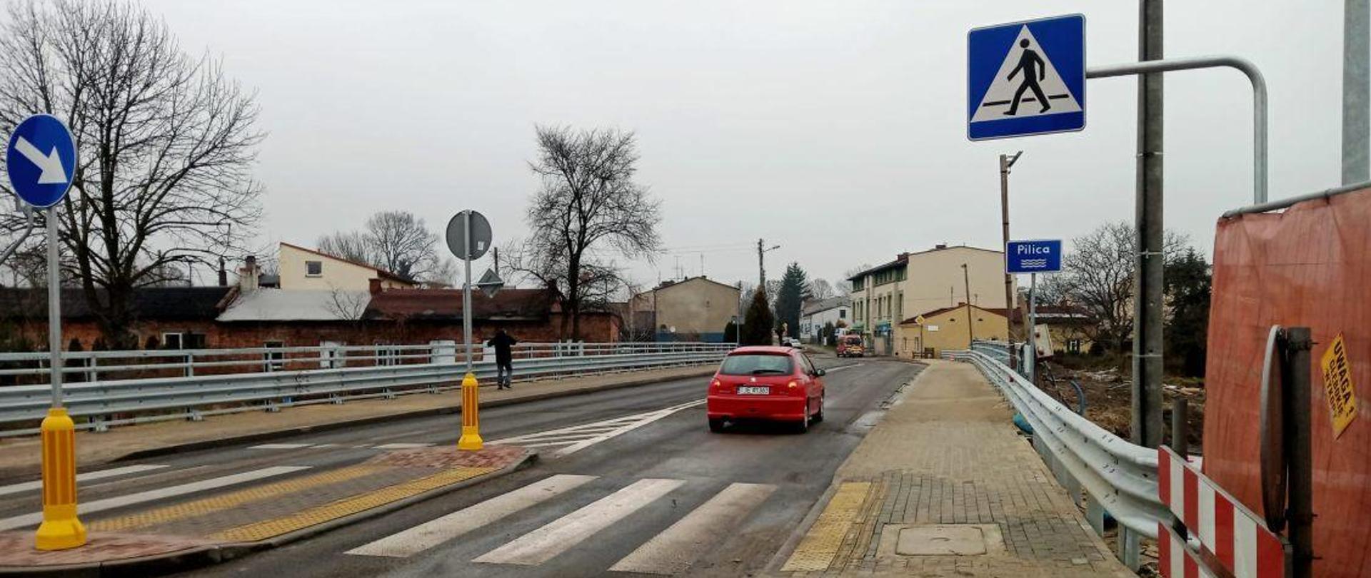Widok nowo wyremontowanego odcinka drogi z przejściem dla pieszych i wyspą dzielącą pasy ruchu. Na jezdni znajduje się czerwony samochód, a po prawej stronie widoczny jest chodnik z barierkami ochronnymi oraz znak informujący o przejściu dla pieszych. W tle znajdują się zabudowania miejscowości Piłka, a po lewej stronie widoczne są dodatkowe znaki drogowe i barierki zabezpieczające.